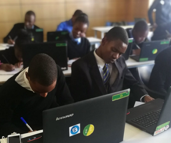 Students in a classroom working on Lenovo laptops while writing notes, demonstrating technology-supported learning and digital education in a school environment.